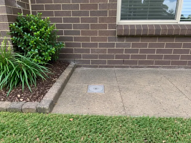 Flush capped access cover in concrete paving beside a brick house wall and garden bed in Seven Hills