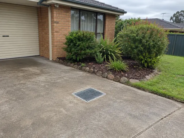 Concrete driveway with a seated stormwater grate beside a brick house and garden in Quakers Hill